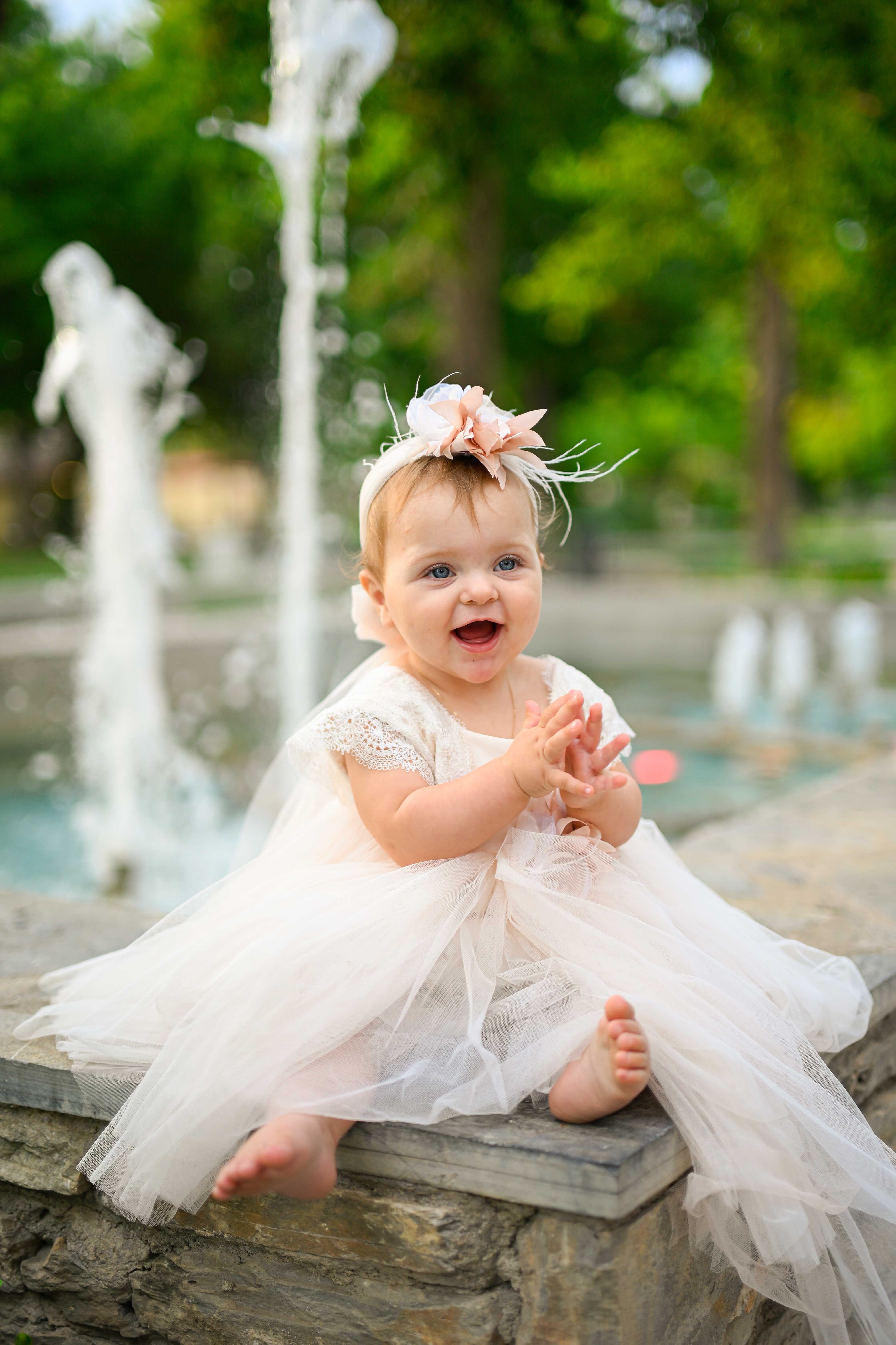 Χαρούμενο μωρό με λευκό φόρεμα βάπτισης δίπλα σε σιντριβάνι - Φωτογραφείο Κολοβός